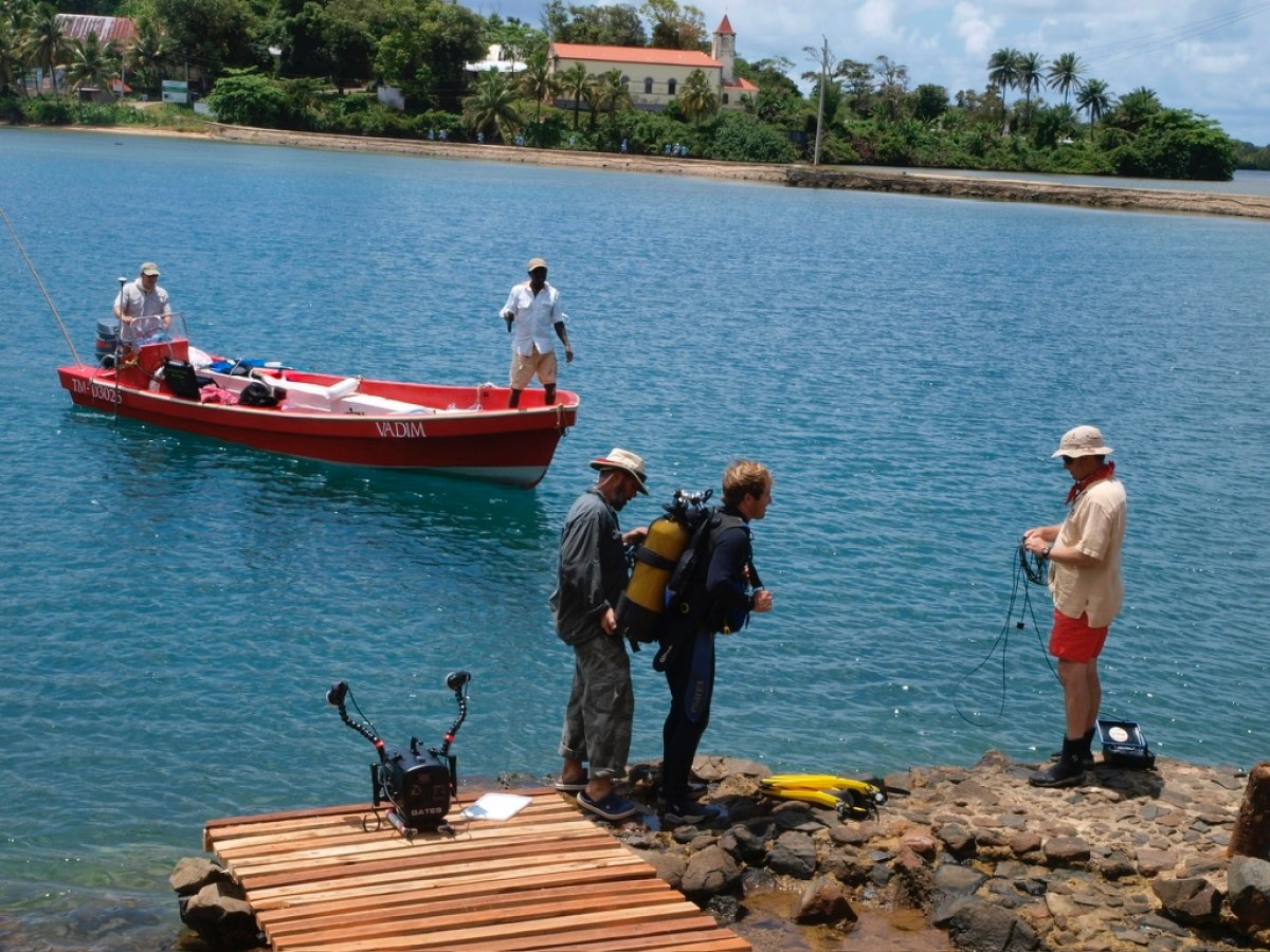 Men on a dock with diving gear, near a red boat on a calm blue bay.