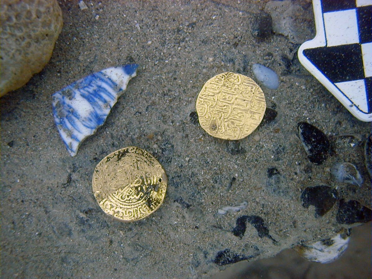 Two gold coins with Arabic script on sand beside ceramic shards.