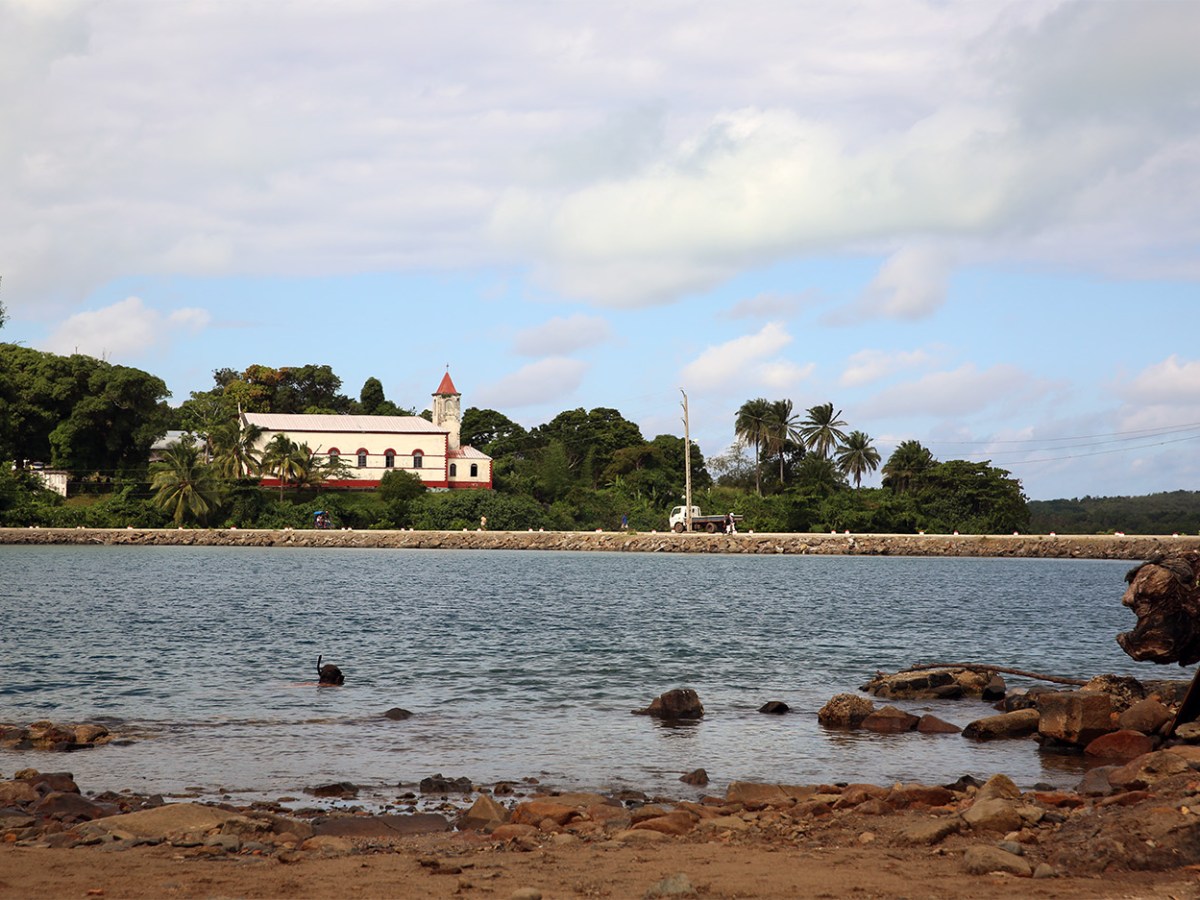 Distant view of a church on a lush island beside calm blue water and a rocky shore under a partly cloudy sky.